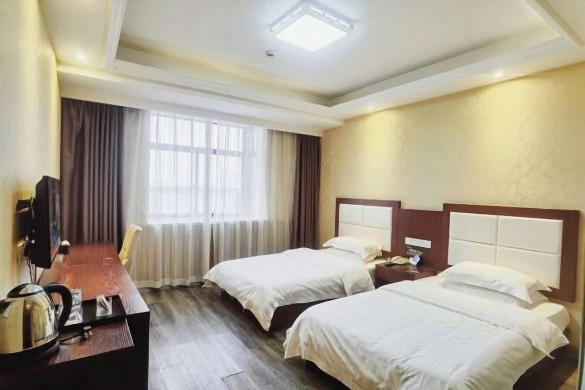 Ein Hotelzimmer mit zwei Betten, einem Schreibtisch mit Wasserkocher, einem Fernseher und einem fenster mit Vorhängen.