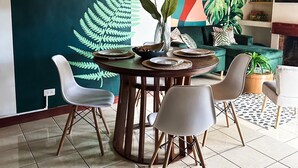 Comedor con una mesa redonda de madera dispuesta para cuatro personas, rodeada de sillas modernas. Se ve una pared verde con diseño de hojas de helecho y un cuadro abstracto colorido.