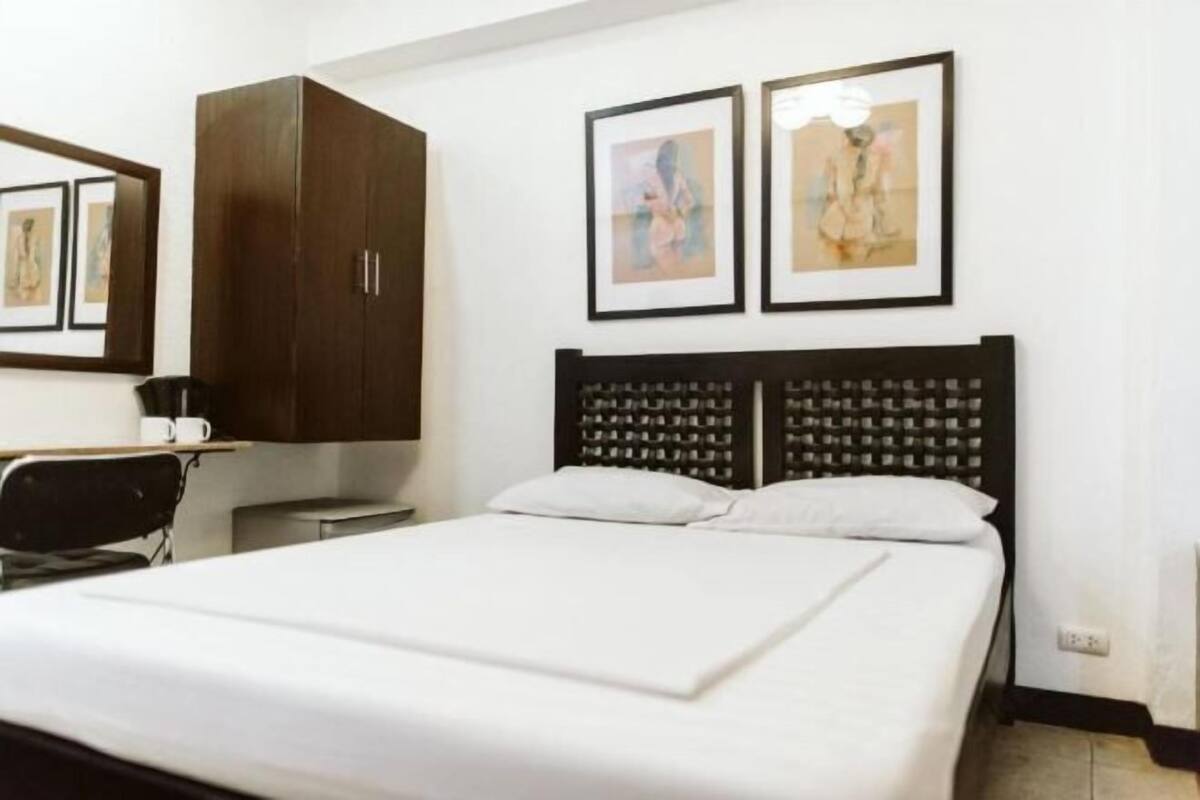 A hotel room with a bed, a desk, a chair, and a wardrobe. There are two framed pictures on the wall above the bed.