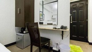 A compact hotel room with a desk, a chair, a small refrigerator, a mirror, and a black door. - San Agustin Residences (Makati)