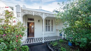 Een wit huis met een zwart-wit geruite inkomhal, een decoratieve veranda en een tuin met diverse planten.
