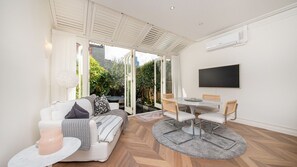 A modern living room with a sofa, a dining table, and chairs. There is a TV mounted on the wall and a window with a view of greenery outside. - Hazel - the perfect escape in Hawksburn (South Yarra)