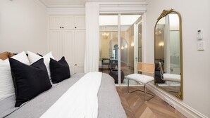 Een slaapkamer met een bed, kussens, een stoel, een spiegel en een deur naar een andere kamer.