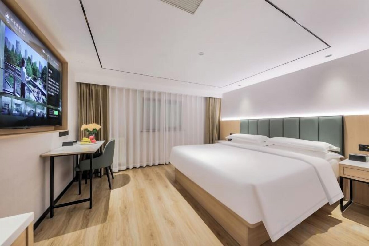 Una habitación de hotel moderna con una cama grande, un escritorio con silla, un televisor y un ventanal con cortinas.