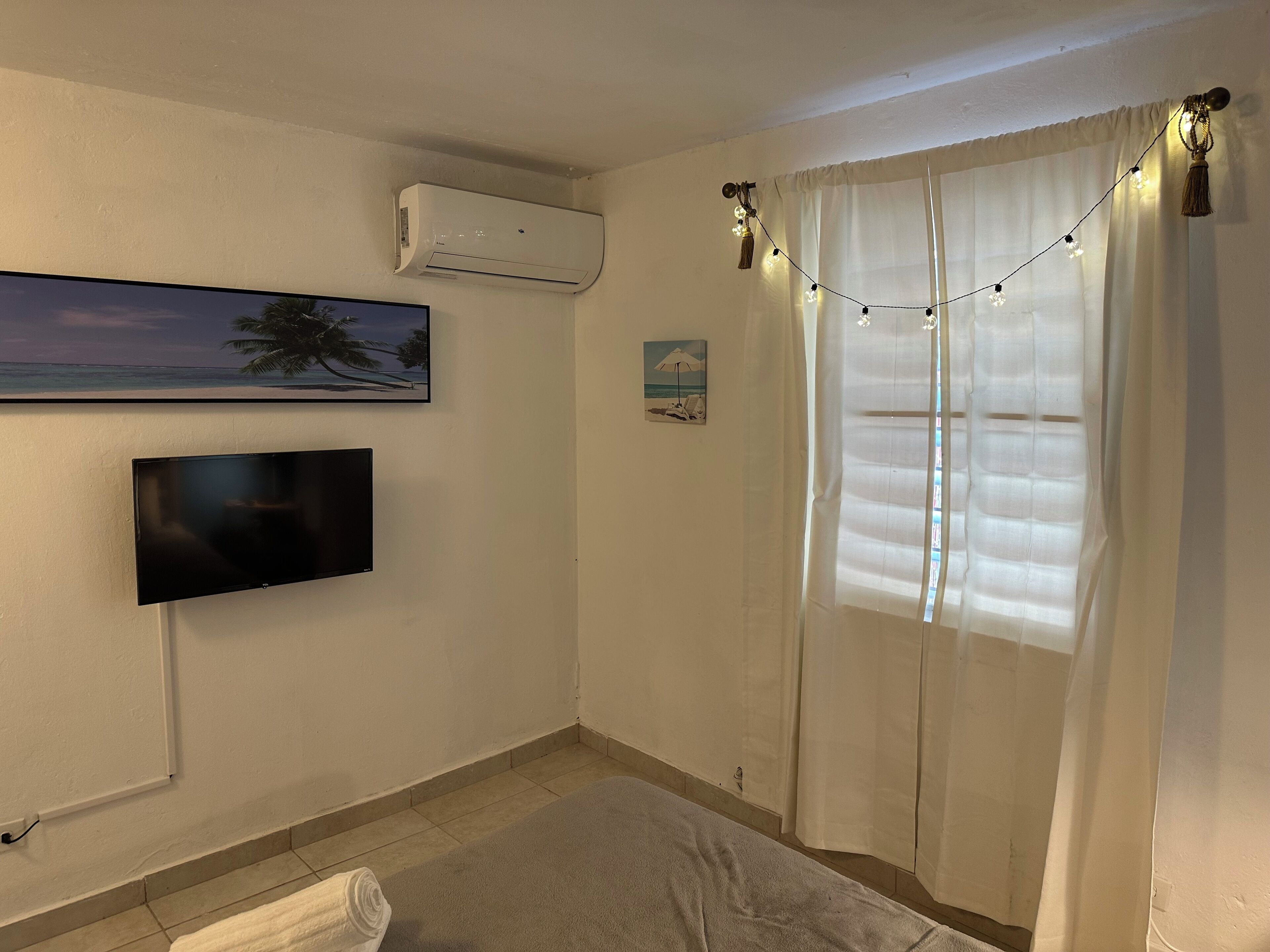 Una habitación con televisión de pantalla plana, aire acondicionado, un cuadro con escena de playa y una ventana con cortinas de gasa y luces colgantes.