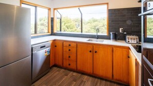 A modern kitchen with wooden cabinets, a stainless steel refrigerator, a dishwasher, a large window, and a view of greenery outside.