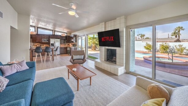 A modern living room with a fireplace, a TV mounted on the wall, and a view of a pool and palm trees through large windows. - Mid Century Modern near STRIP w Pool King Suite (Las Vegas)