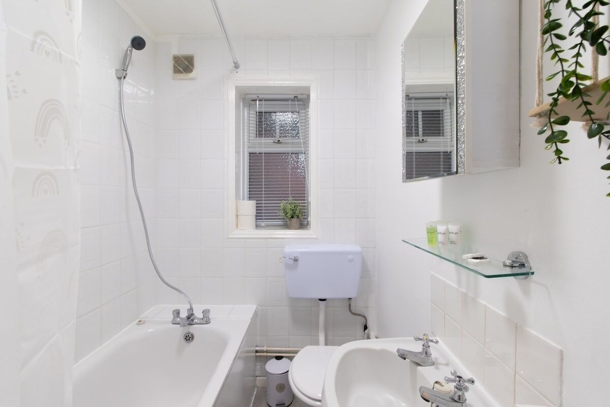 Ein modernes Badezimmer mit einer Badewanne, Toilette und Waschbecken. Ein Fenster mit Jalousien und ein kleiner Topf mit Pflanze auf der Fensterbank.