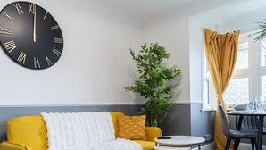 A room with a yellow sofa, a white furry throw blanket, a round table with chairs, a large wall clock, and a potted plant. - Open Mind Property - Cosy Apartment with 3 Beds (Portsmouth)