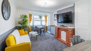 A modern living room with a yellow sofa, a fireplace, a dining area, and a large clock on the wall. - Open Mind Property - Cosy Apartment with 3 Beds (Portsmouth)