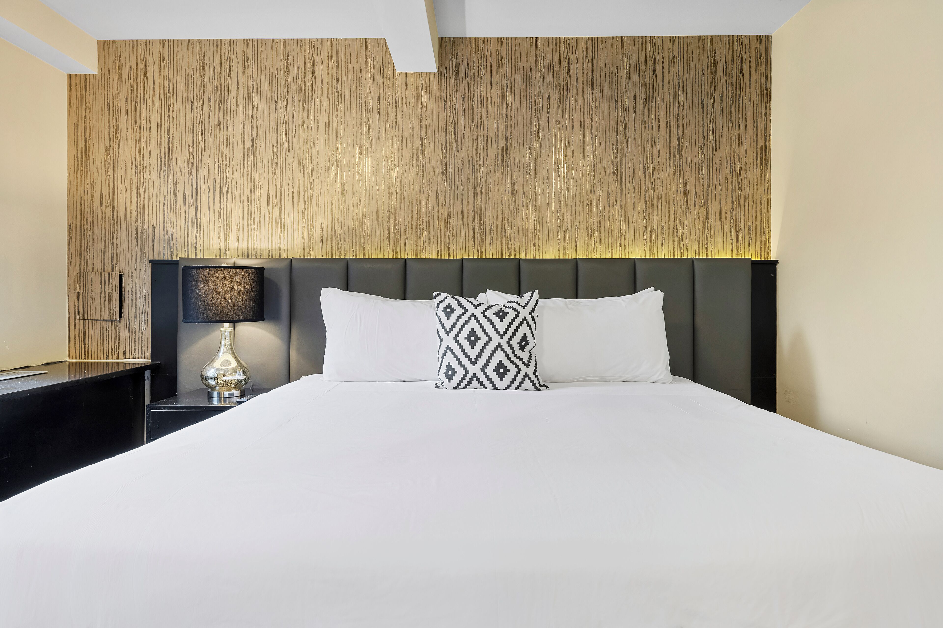 Quarto de hotel com uma cama grande, uma mesa de cabeceira com abajur e uma parede texturizada.
