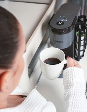 A person holding a white mug with coffee, a Keurig coffee maker, and a black coffee pod holder on a white tray.
