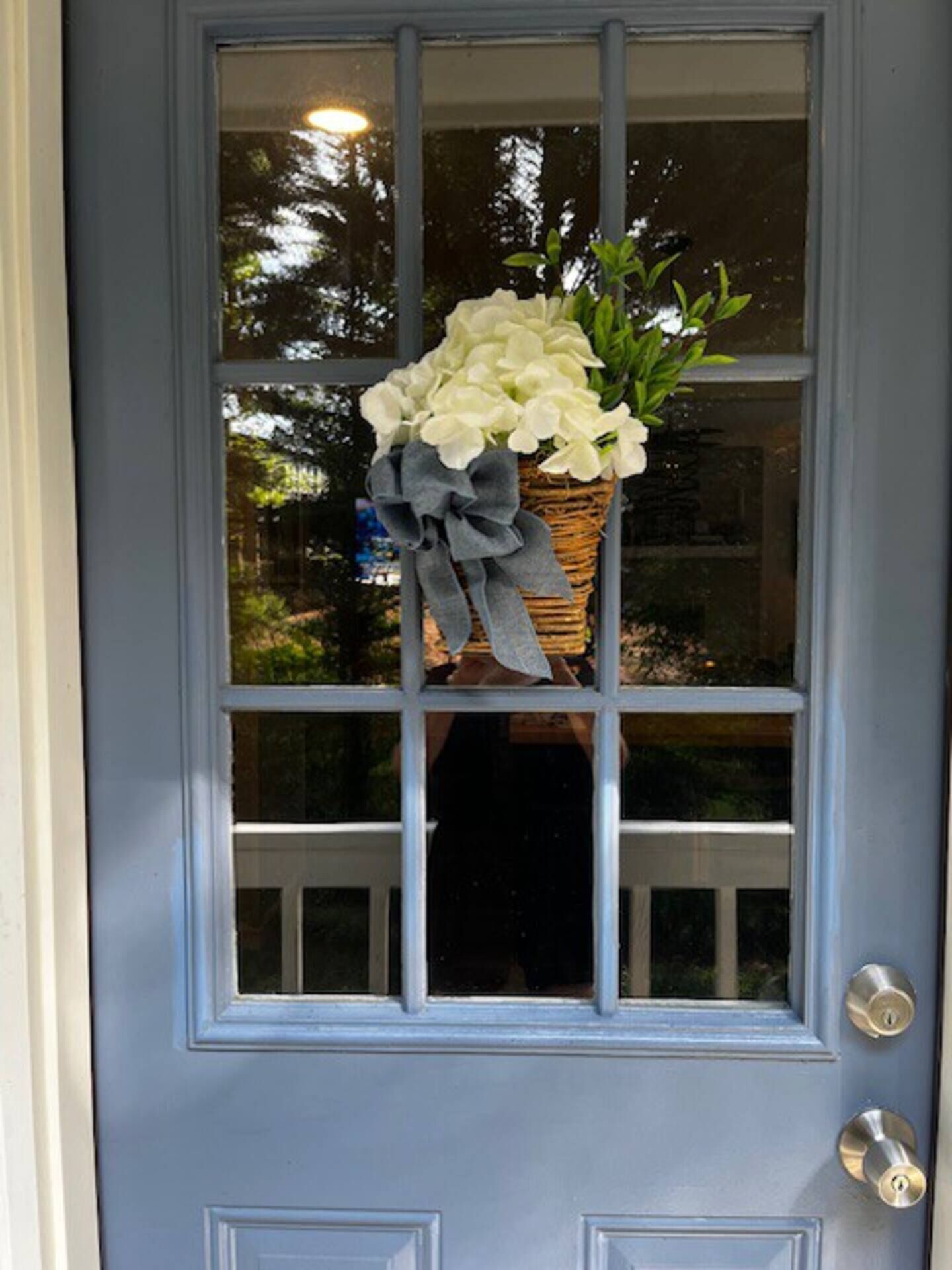 Eine blaue Tür mit einem Fenster, ein Korb voller weißer Blumen und ein graues Band.