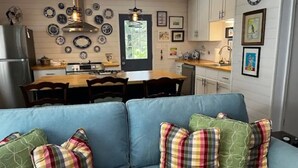 A modern kitchen with a dining area, featuring a blue sofa with patterned cushions, a stainless steel refrigerator, and a variety of decorative plates on the wall.