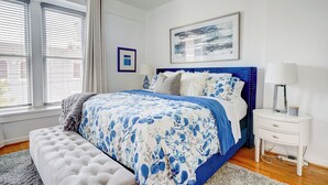 A bedroom with a large bed, a tufted bench, a nightstand, and a ceiling fan. - The Art Loft - A Luxurious 2-BdRm in the Heart of Historic Downtown Wilmington (Wilmington)