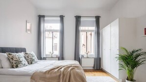 Une chambre à coucher avec un lit, deux fenêtres avec des rideaux, un radiateur, une plante en pot et une armoire.