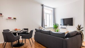 Un salon moderne avec un canapé gris, une table à manger, des chaises, un téléviseur à écran plat et une fenêtre avec des rideaux.