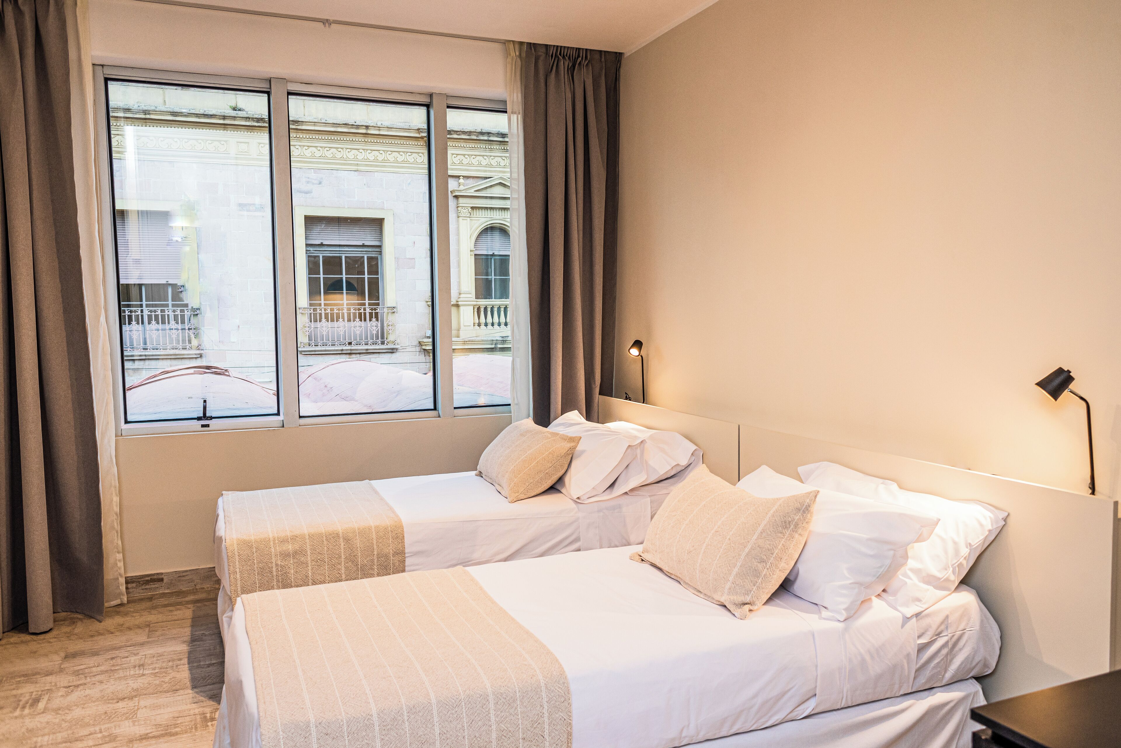 Quarto de hotel com duas camas, uma janela grande com cortinas e vista para um edifício.