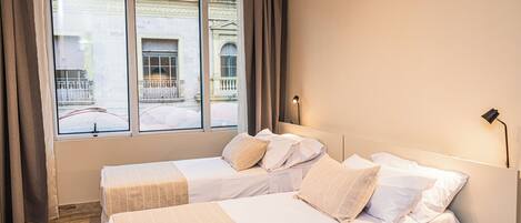 Quarto de hotel com duas camas, uma janela grande com cortinas e vista para um edifício.
