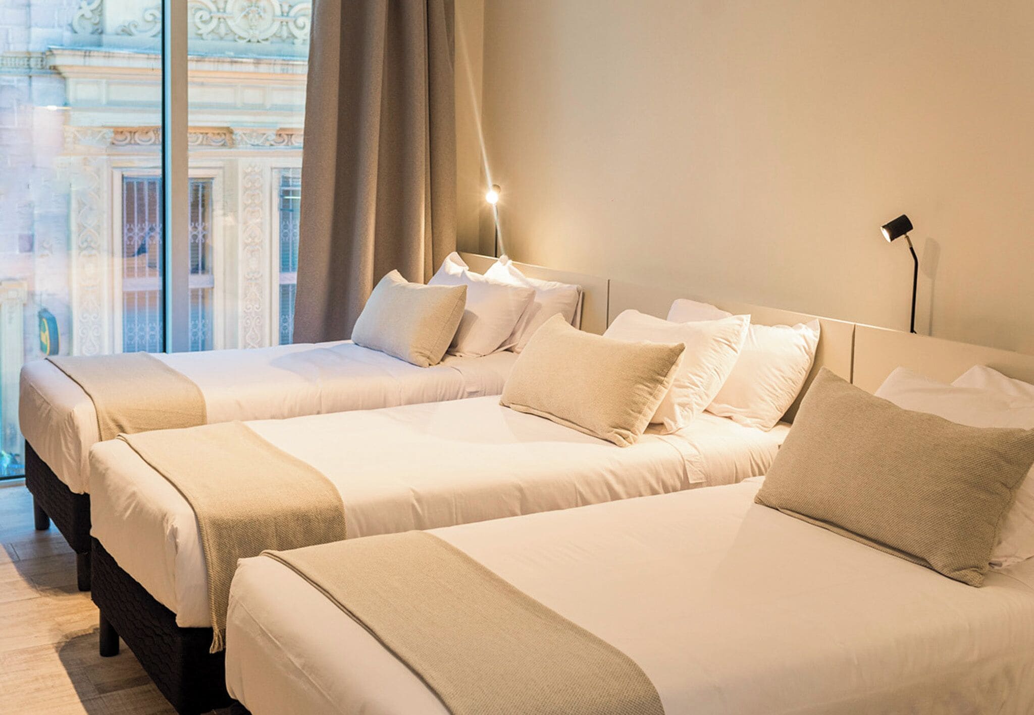 A hotel room with two beds, a large window with a view of a building, and a bedside lamp.
