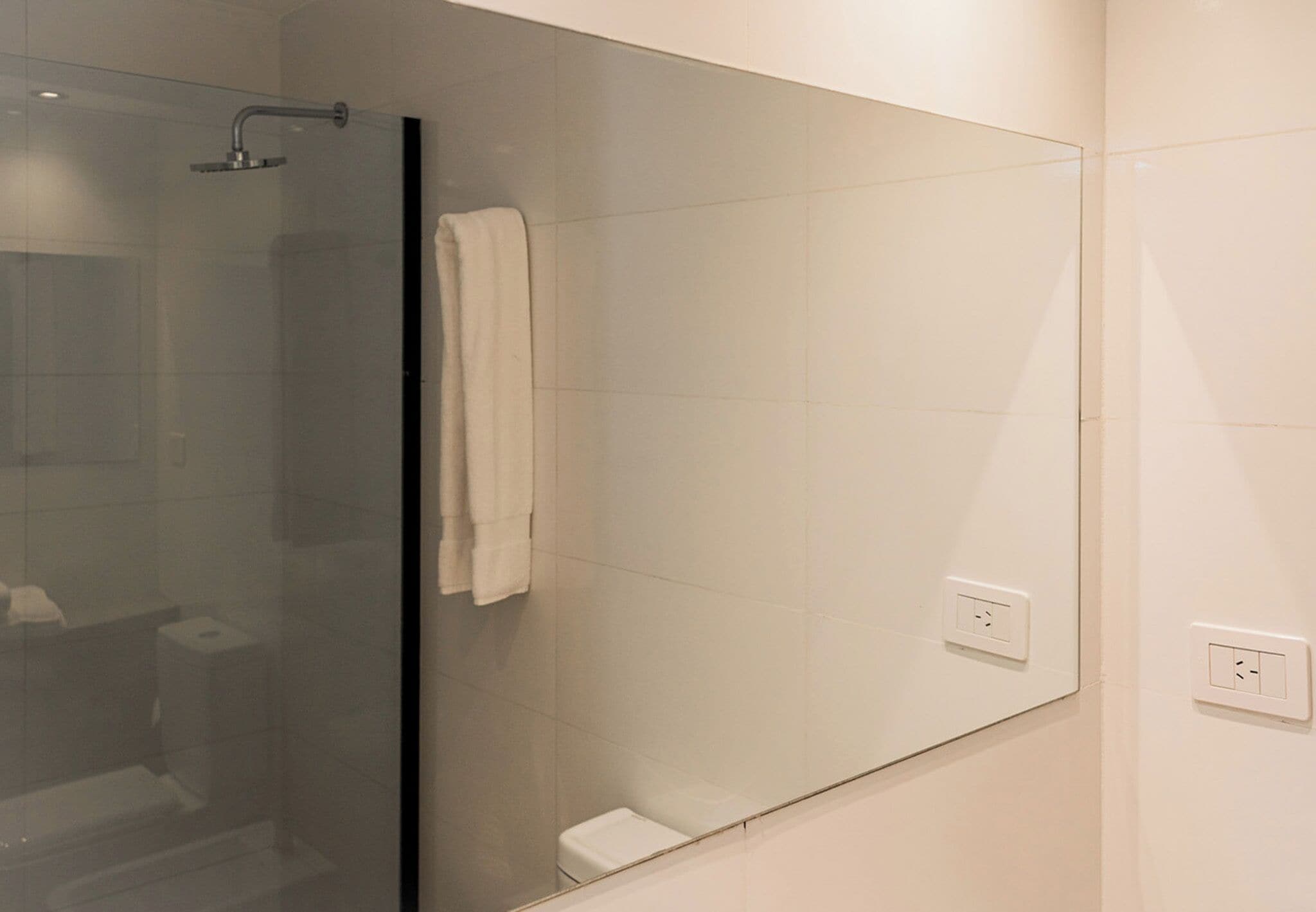 A modern bathroom with a glass shower enclosure, a white towel hanging on the wall, and a visible electrical outlet.