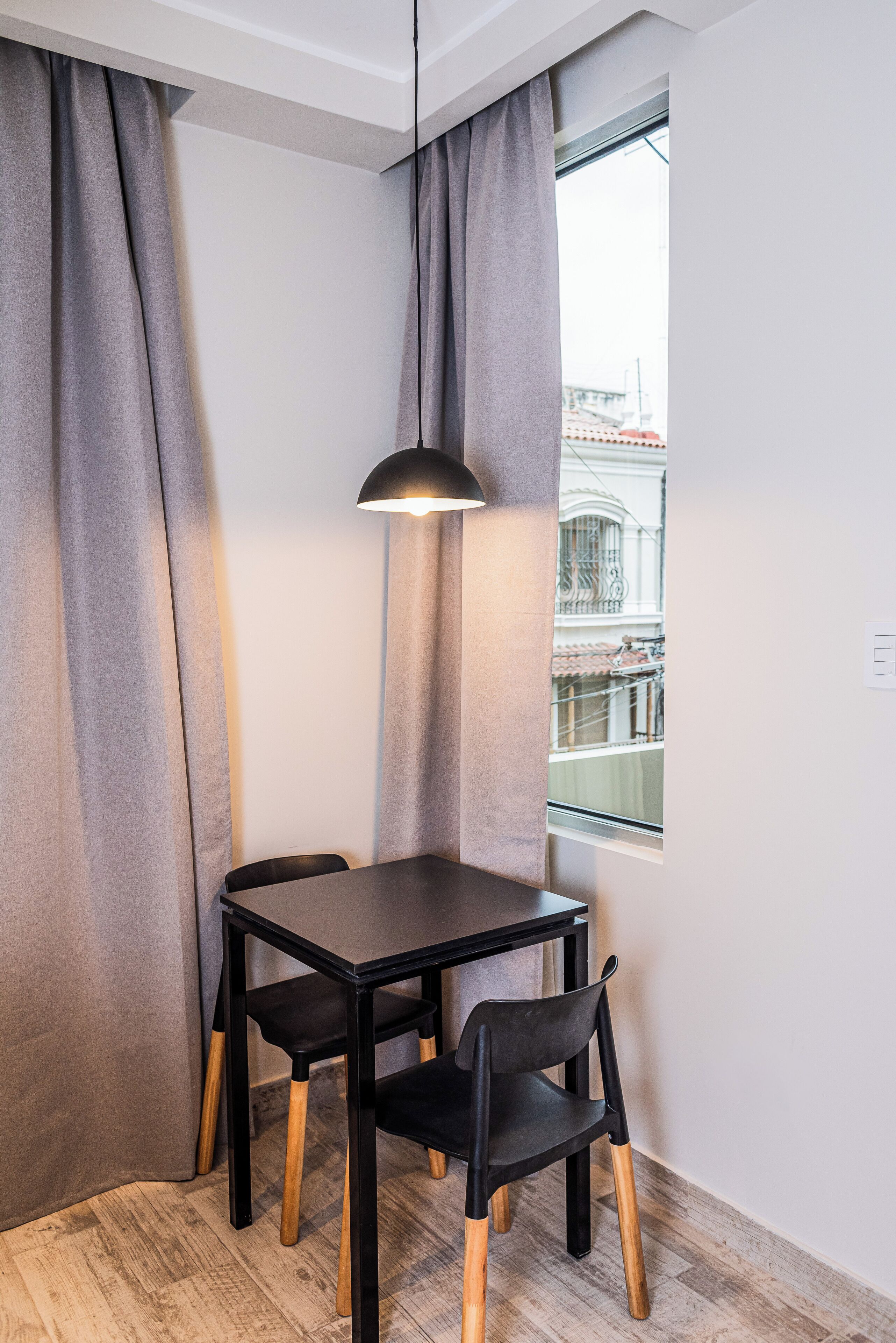 A corner room with a table and two chairs, a hanging light, and a window with a view of buildings.