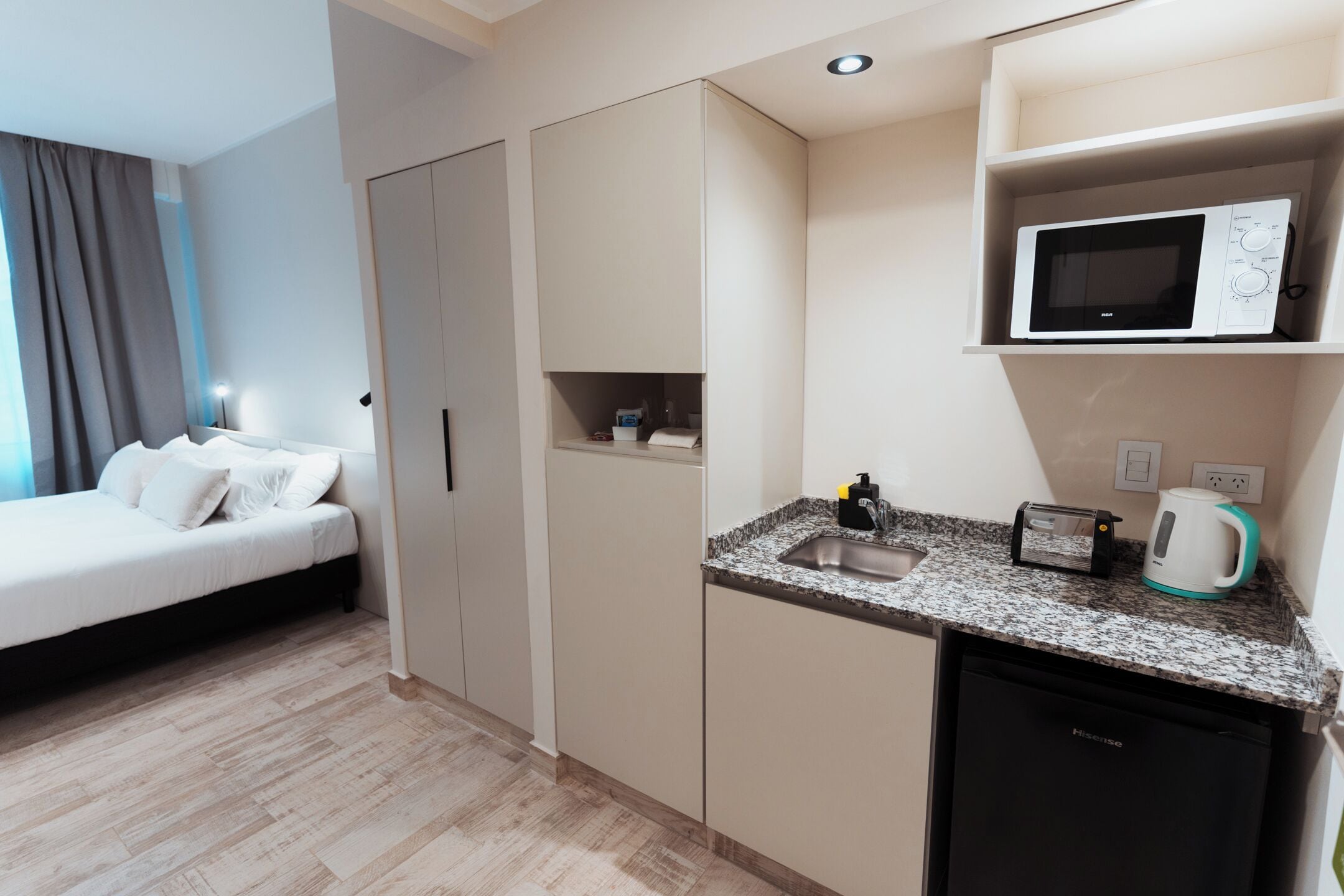 A compact hotel room with a bed, kitchenette, and a small refrigerator.