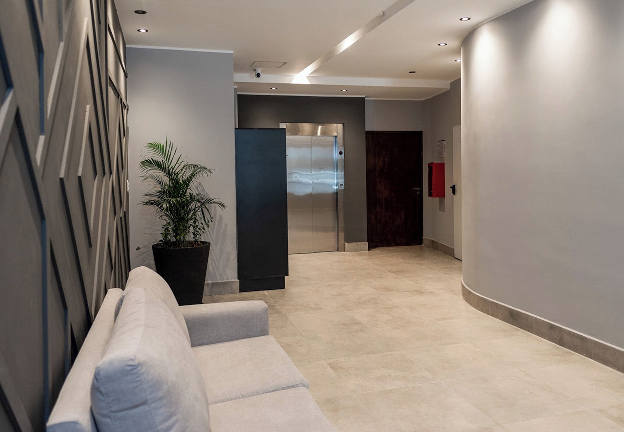 A modern lobby with a seating area, a large potted plant, a glass elevator, and a red fire extinguisher.