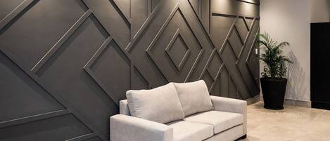 Un intérieur moderne avec un canapé gris placé devant un mur au motif géométrique, une plante en pot et un sol carrelé.