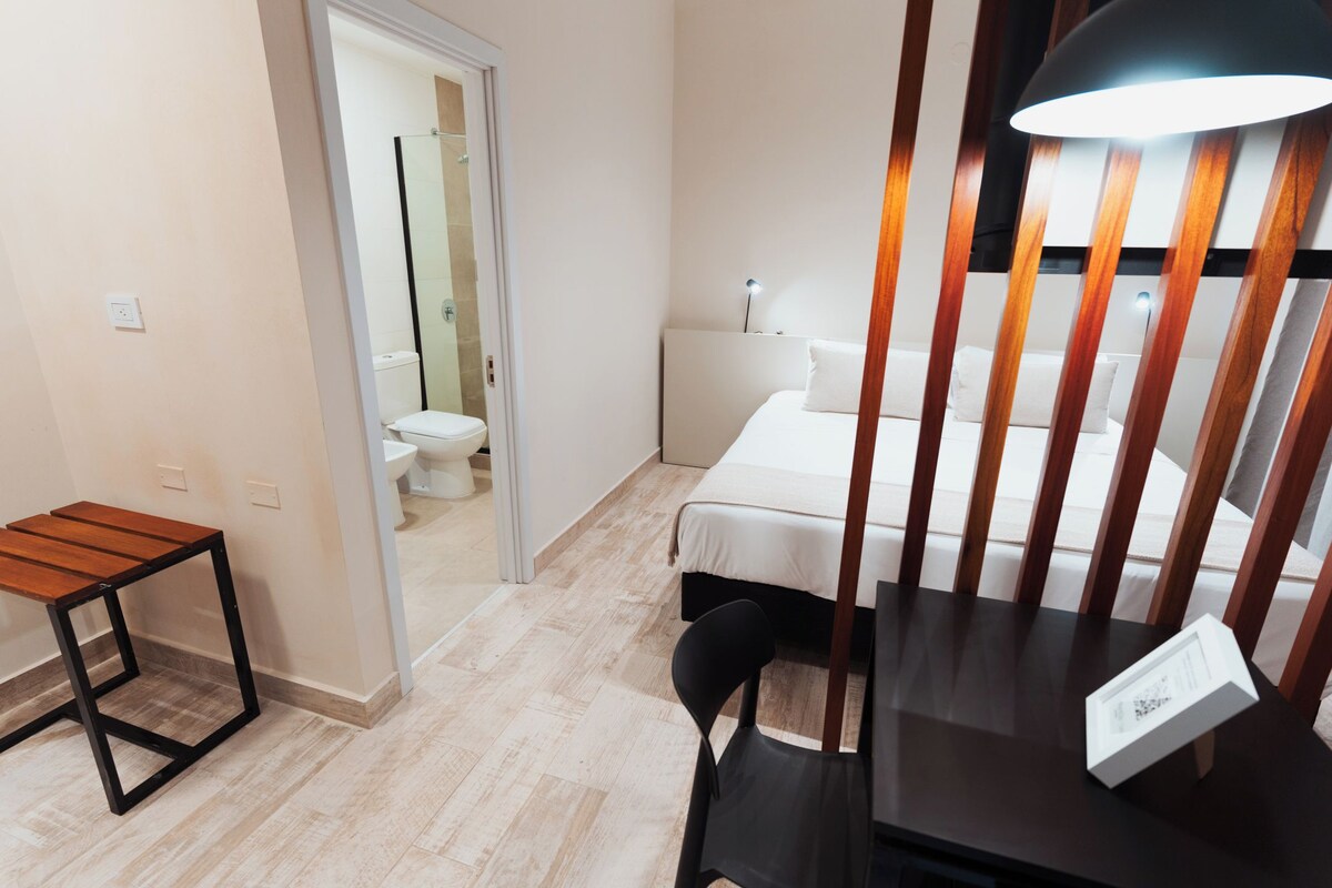 A small, modern room with a bed, a desk, and a chair. There is a wooden table and a lamp on the desk.
