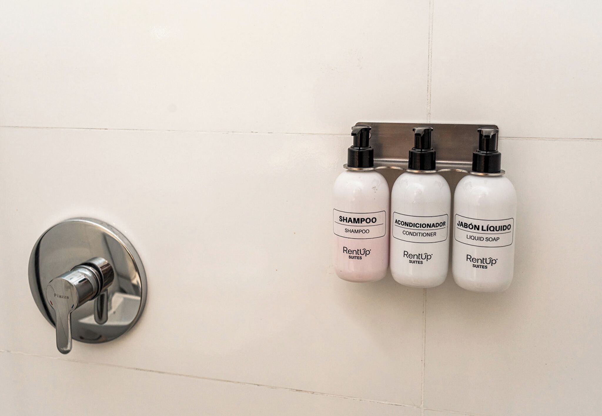 A bathroom wall with a mounted soap dispenser and three bottles of shampoo, conditioner, and liquid soap.