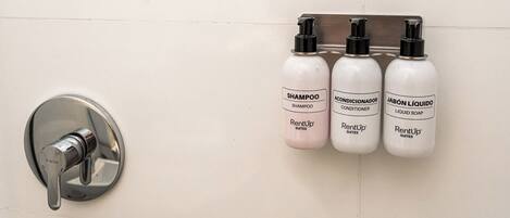 A bathroom wall with a mounted soap dispenser and three bottles of shampoo, conditioner, and liquid soap.