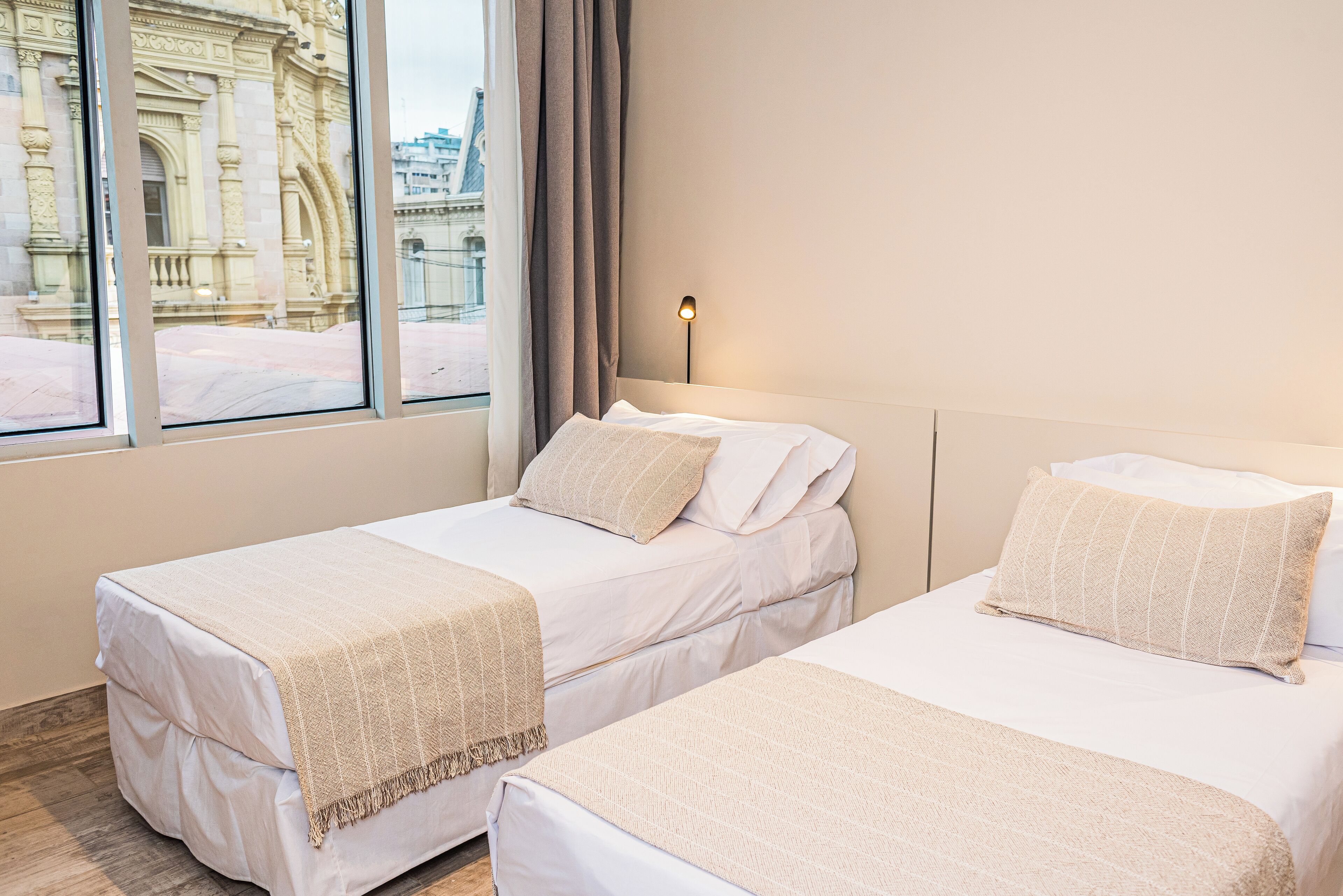 Two single beds with white linens and beige throw blankets, a nightstand with a lamp, and a view of a historic building through a window with curtains.