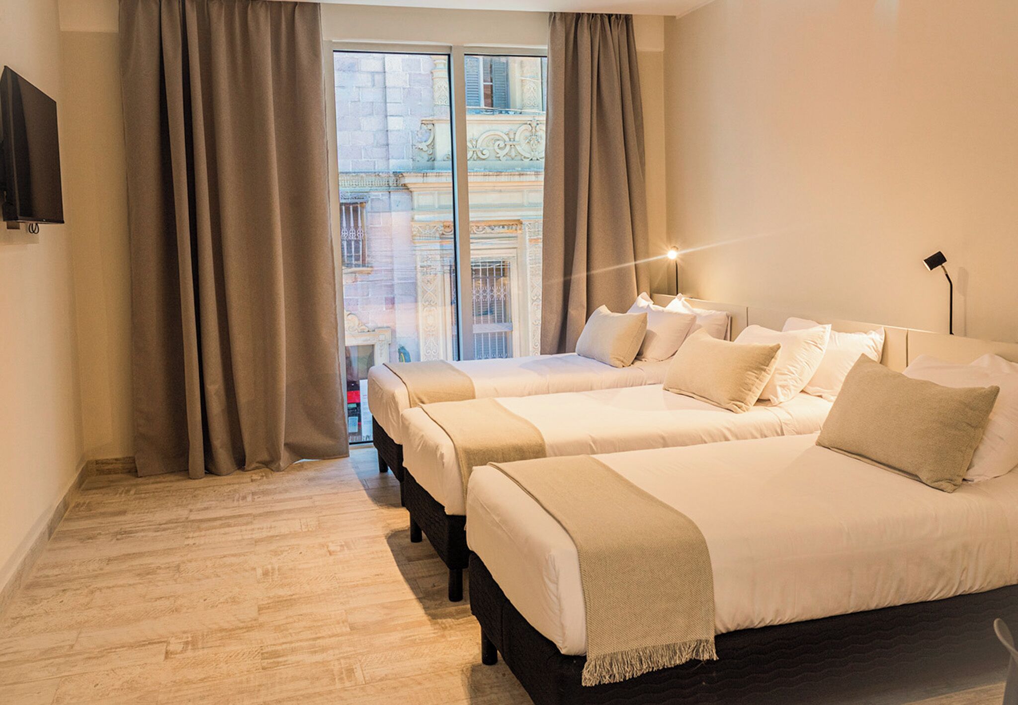 A hotel room with two beds, a TV, a window with curtains, and a view of a building.