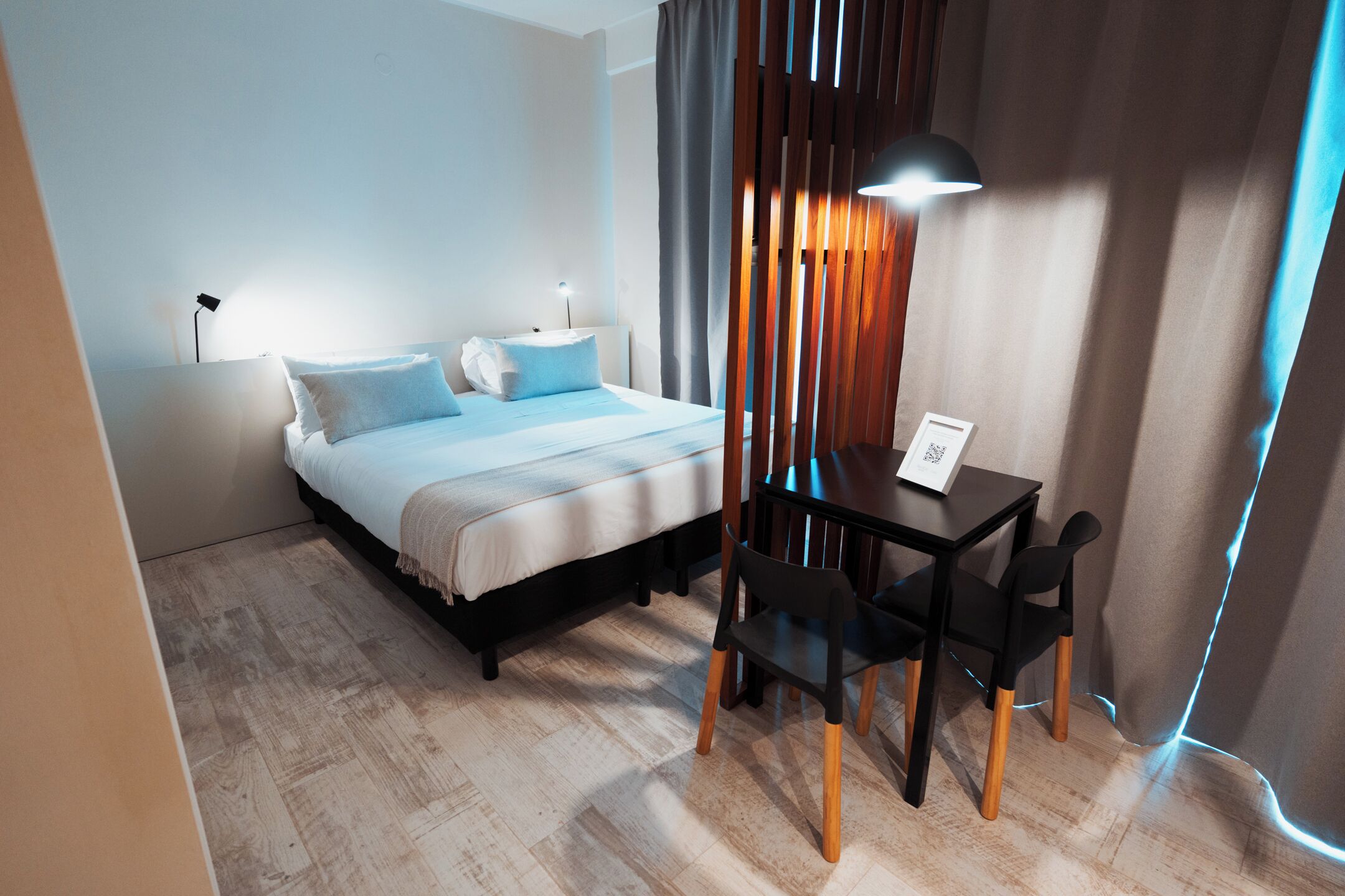 A modern hotel room with a bed, bedside table, lamp, and a desk with chairs.