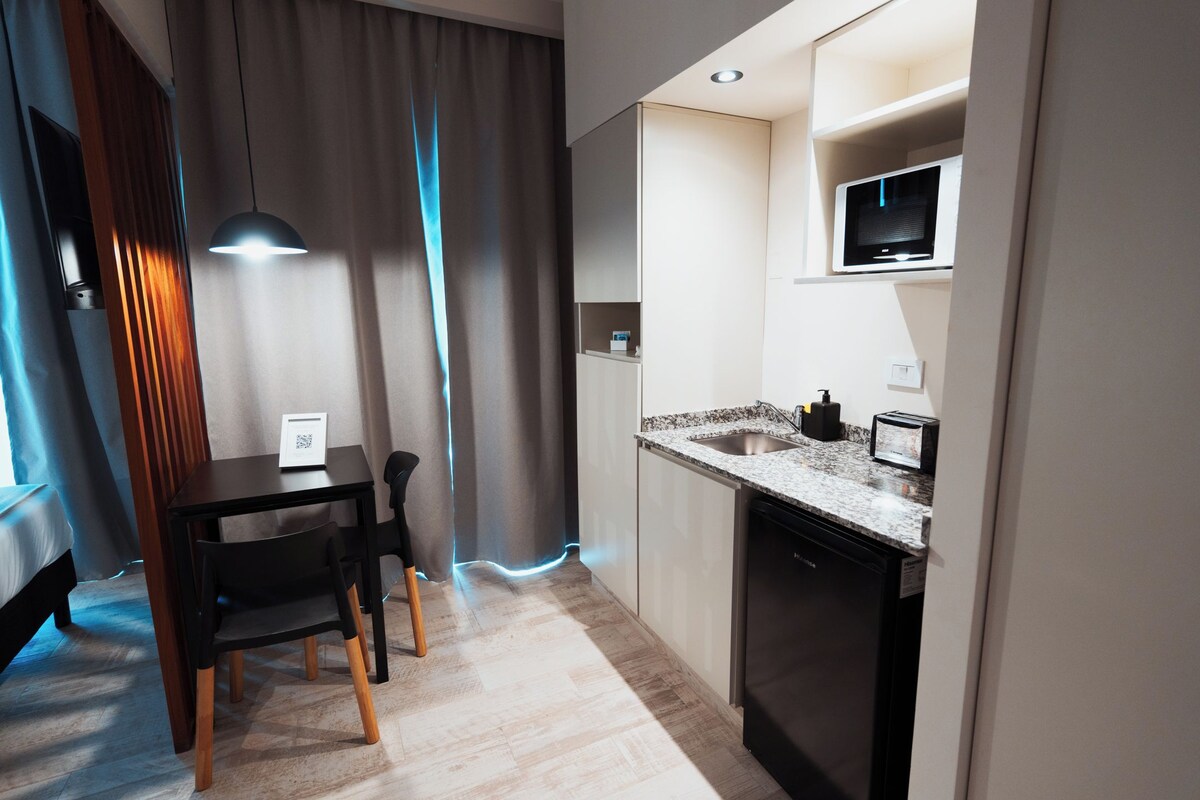 Uma cozinha moderna com bancada de granito, micro-ondas embutido e lava-louças. Há uma área de jantar com mesa e cadeiras, e uma luminária pendente sobre a mesa.