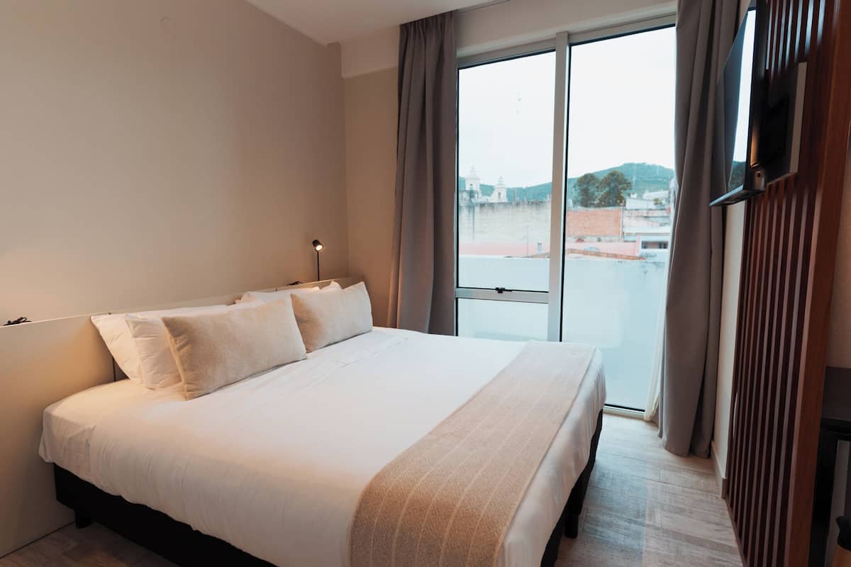 Een hotelkamer met een groot bed, een aan de muur bevestigde televisie en uitzicht op een stadsgezicht door het raam.