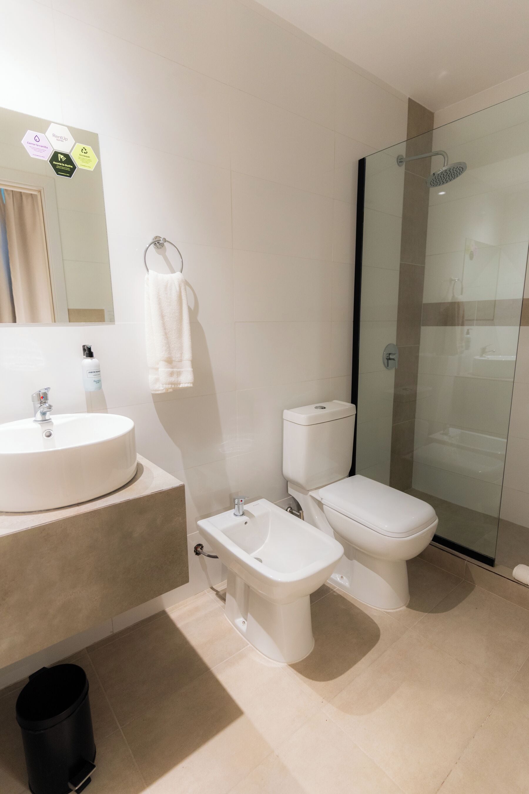 Une salle de bain moderne avec un lavabo rond, deux toilettes, un bidet, une douche à parois de verre et un miroir sur lequel est accrochée une serviette.