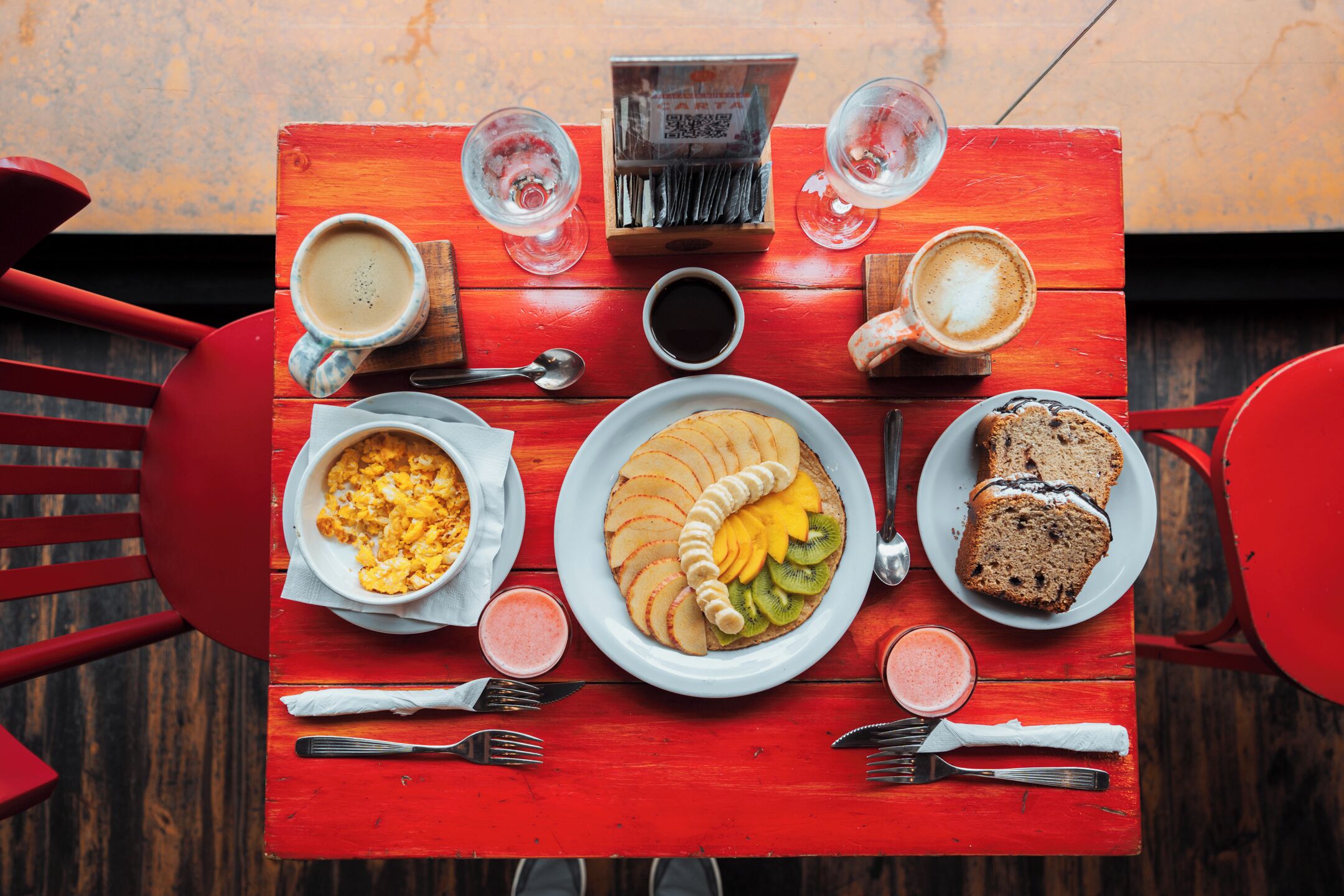 A red wooden table with breakfast items including eggs, fruit, and bread, along with coffee and a menu.