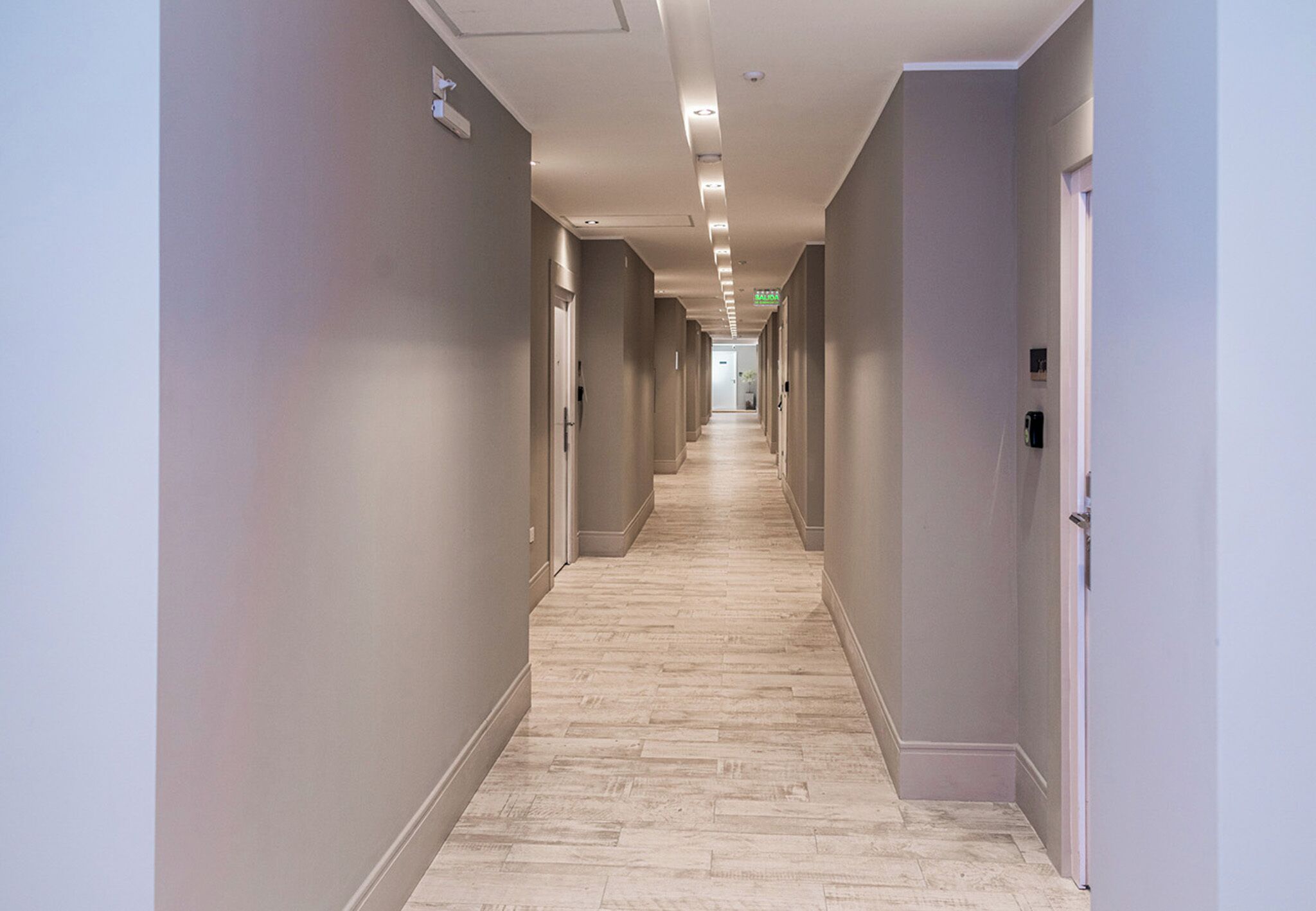 A long, narrow hallway with a light-colored wooden floor and walls painted in a neutral tone.