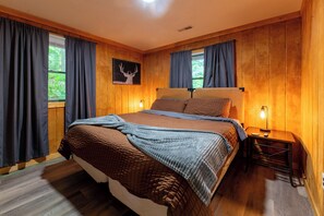 A wooden-paneled bedroom with a bed, curtains, a nightstand, and a wall-mounted picture.
