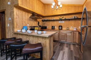 A wooden kitchen with a central island, bar stools, a stainless steel refrigerator, and a sign reading 'KITCHEN.'