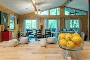 A wooden interior with a dining table, chairs, a glass bowl of lemons, and a clock on the wall.
