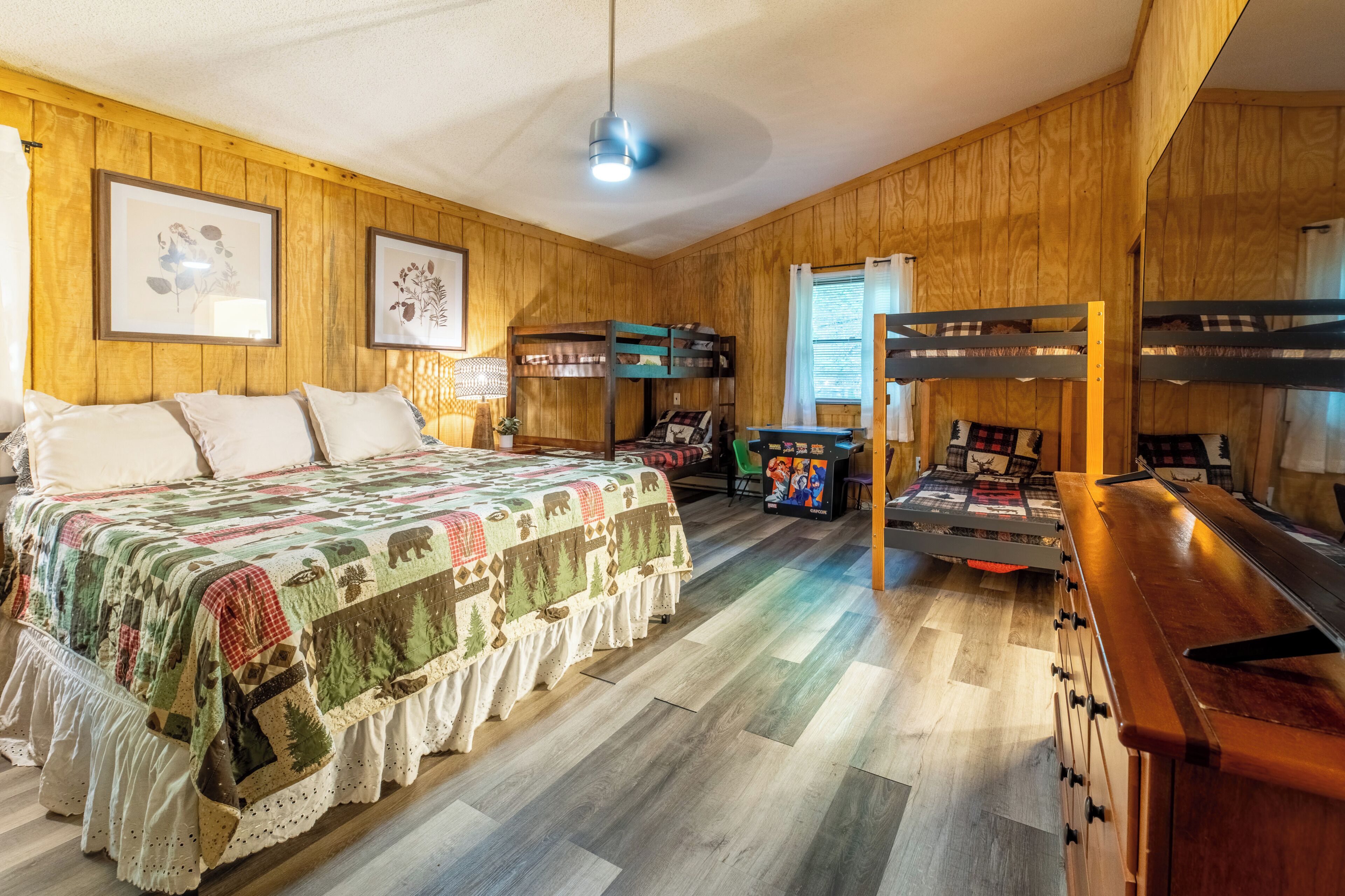 Une chambre avec un lit superposé, un grand lit recouvert d’une courtepointe, des murs en bois, un ventilateur de plafond et une fenêtre avec des rideaux.