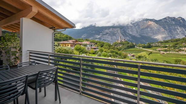A balcony with a view of a mountainous landscape, a table with chairs, and a building with a red-tiled roof.