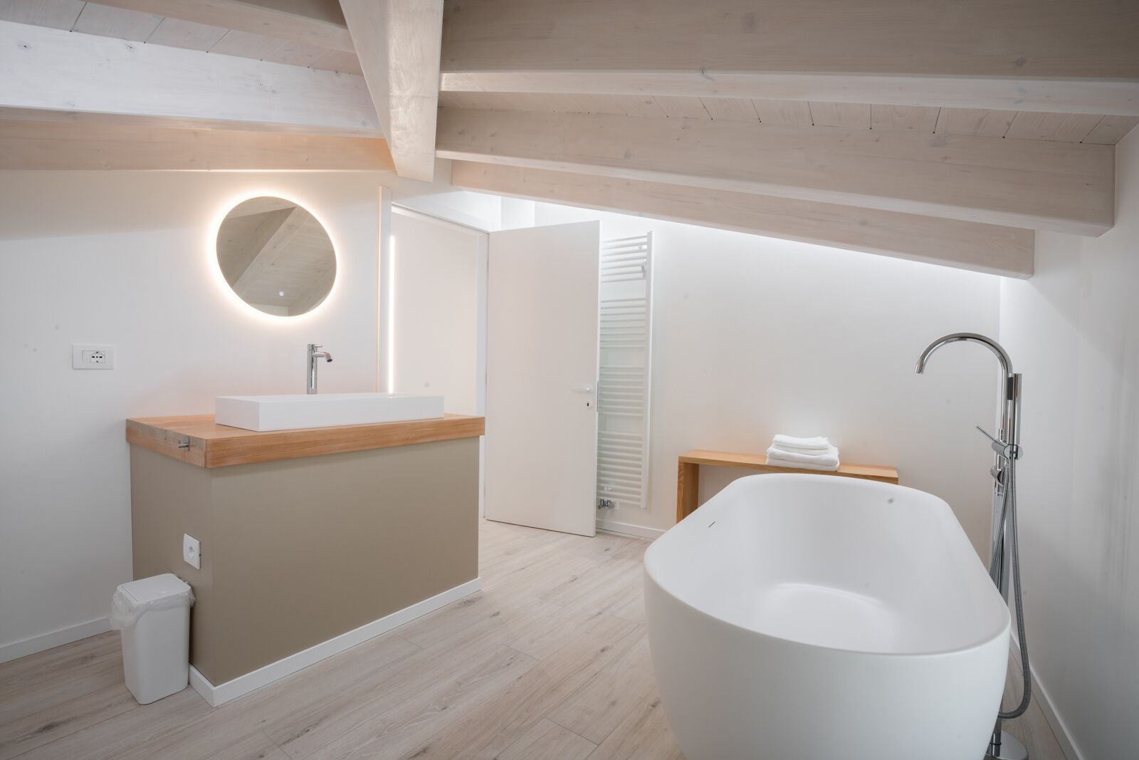 Une salle de bain moderne avec une baignoire sur pieds, un grand miroir rond doté d’un éclairage intégré, un meuble-lavabo en bois et une douche à l’italienne.