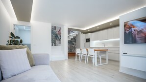A modern living room with a sofa, a dining area with chairs, a kitchen with white cabinets, and a flat-screen TV mounted on the wall.