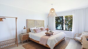 A bedroom with a large bed, a bedside table with a lamp, a wooden chair, a window with a view of trees, and a hanging light fixture. - Waterfront Getaway, Kayaks included in stay - Kid Friendly (Sanctuary Point)