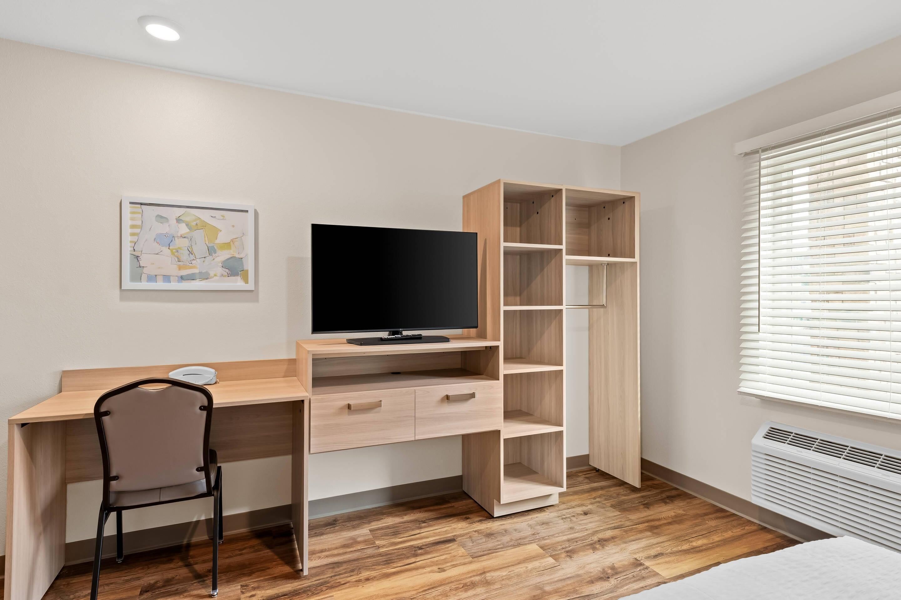 A modern bedroom with a wooden desk, a TV, a chair, and a large window with blinds.