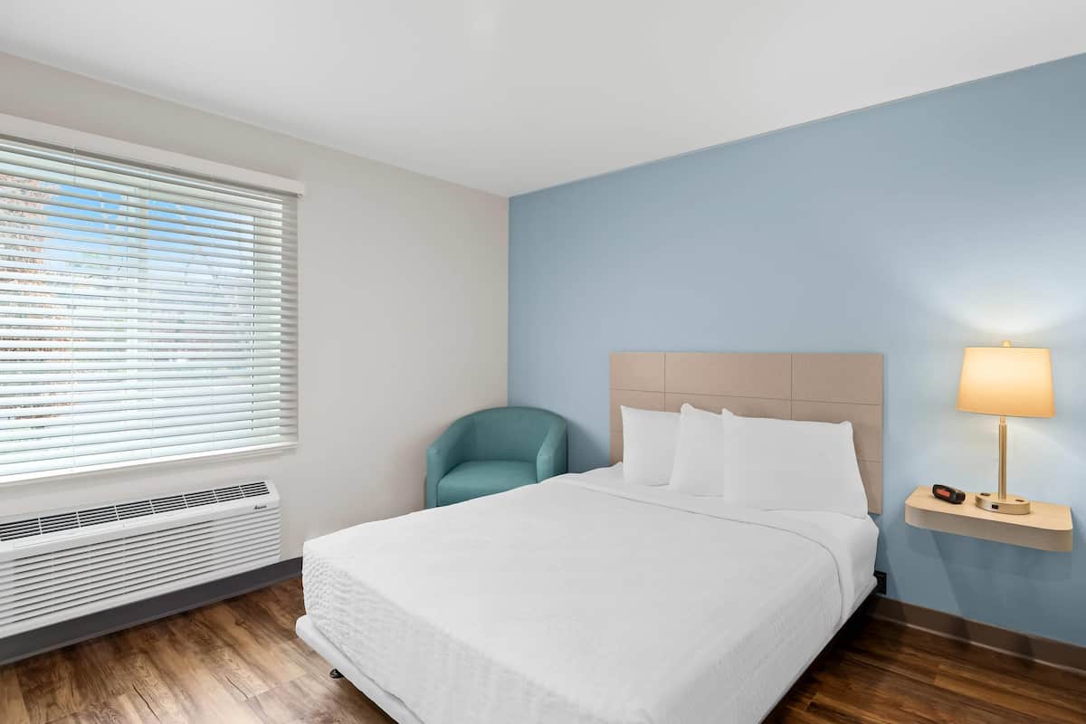 Una habitación de hotel con una cama, una silla, una mesita de noche con lámpara y una ventana con persianas.